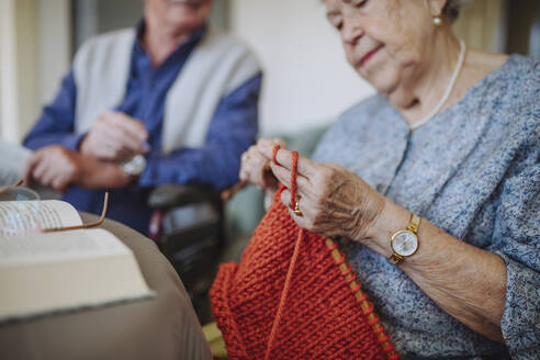 Ältere Frau, die Wolle strickt, mit einem sitzenden Mann im Hintergrund - HAPF03269