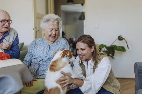 Happy senior couple with healthcare worker stroking dog at home - HAPF03274