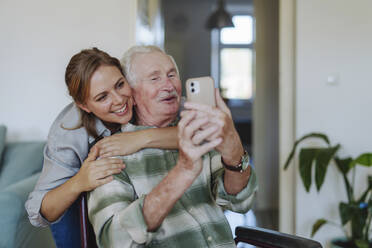 Smiling healthcare worker hugging senior man taking selfie at home - HAPF03283