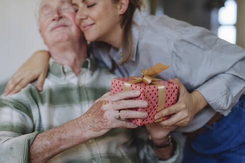 Smiling healthcare worker embracing and giving gift to senior man on birthday - HAPF03289