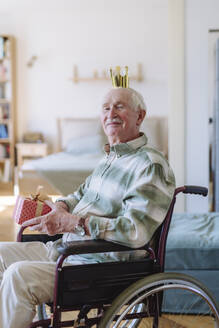 Lächelnder älterer Mann, der eine Krone trägt und ein Geschenkpaket zu Hause in der Hand hält - HAPF03290