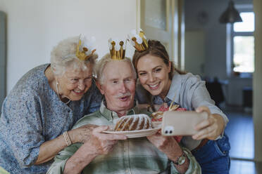 Happy healthcare worker taking selfie with senior couple holding cake and celebrating birthday at home - HAPF03292