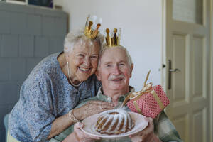Glückliche ältere Frau mit Mann hält Geburtstagskuchen zu Hause - HAPF03294