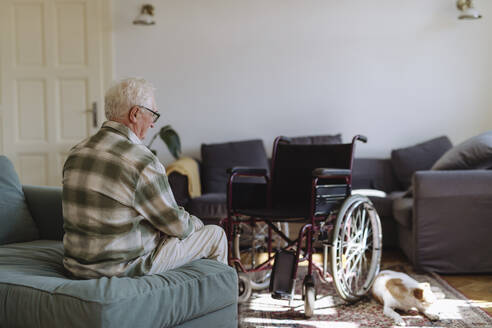 Trauriger älterer Mann sitzt auf dem Sofa in der Nähe von Rollstuhl im Wohnzimmer zu Hause - HAPF03311