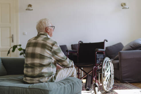 Nachdenklicher älterer Mann sitzt auf dem Sofa in der Nähe des Rollstuhls zu Hause - HAPF03313