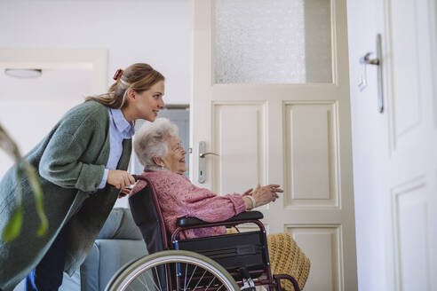 Lächelnde Pflegekraft schiebt den Rollstuhl einer älteren Frau zu Hause - HAPF03325