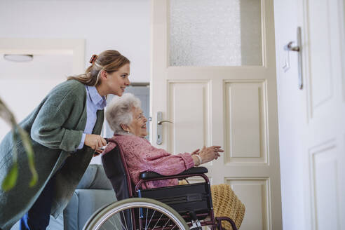 Smiling healthcare worker pushing senior woman's wheelchair at home - HAPF03325