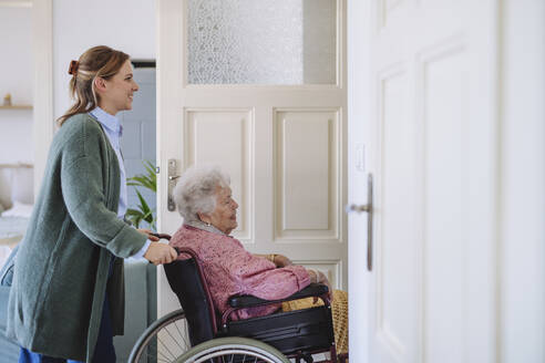 Happy healthcare worker pushing senior woman's wheelchair at home - HAPF03326