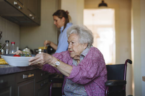 Senior woman holding bowl with home caregiver working in background - HAPF03329
