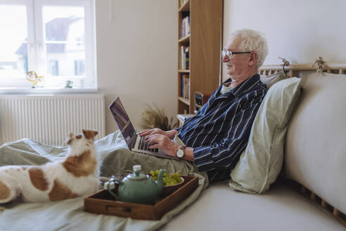 Senior man using laptop with dog sitting on bed at home - HAPF03341