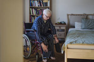 Älterer Mann sitzt und stellt den Rollstuhl in der Nähe des Bettes im Schlafzimmer zu Hause ein - HAPF03346