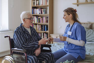 Lächelnde Pflegekraft, die einem im Rollstuhl sitzenden älteren Mann Medikamente verabreicht - HAPF03350