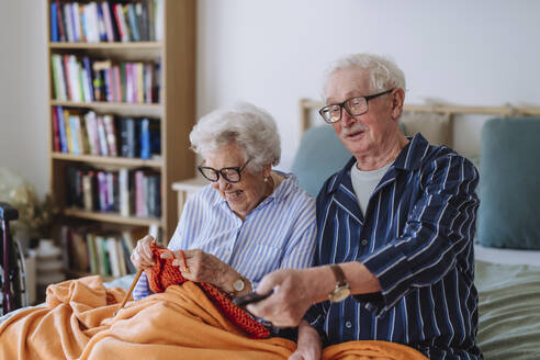 Glückliche ältere Frau strickt mit Mann, der die Fernbedienung hält und zu Hause fernsieht - HAPF03366