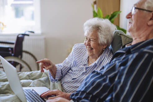 Happy senior woman pointing over laptop with man sitting on bed at home - HAPF03370