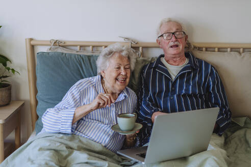 Glücklicher älterer Mann und Frau mit Laptop auf dem Bett zu Hause - HAPF03372