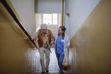 Glückliche Pflegekraft, die mit einem älteren Mann auf einer Treppe zu Hause nach oben geht - HAPF03379