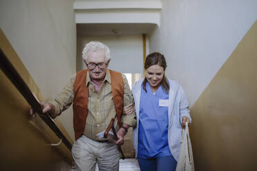 Lächelnde Pflegekraft, die mit einem älteren Mann auf einer Treppe zu Hause aufsteigt - HAPF03380
