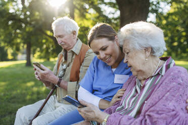 Lächelndes Pflegepersonal mit einem älteren Mann und einer Frau, die ein Smartphone im Park benutzen - HAPF03381