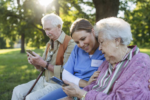 Smiling healthcare worker with senior man and woman using smart phone in park - HAPF03381