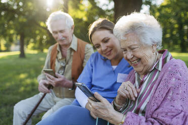 Glückliche Mitarbeiterin im Gesundheitswesen mit älterem Mann und Frau, die ein Smartphone im Park benutzen - HAPF03382
