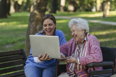Glückliche Mitarbeiterin im Gesundheitswesen mit älterer Frau, die einen Laptop im Park benutzt - HAPF03386