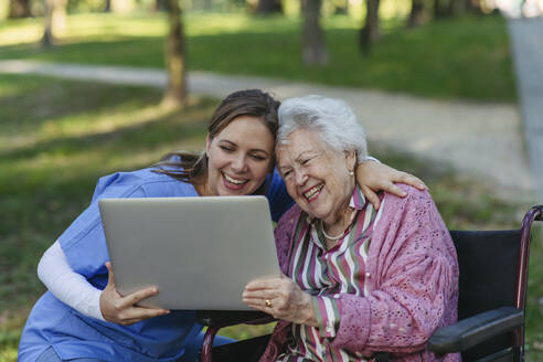 Lächelnde Krankenschwester mit älterer Frau, die im Park einen Laptop benutzt - HAPF03387