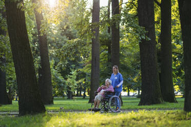 Lächelndes Pflegepersonal geht mit einer älteren Frau im Rollstuhl in der Nähe von Bäumen im Park spazieren - HAPF03394