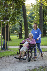 Glückliche Mitarbeiterin im Gesundheitswesen geht mit einer älteren Frau im Rollstuhl im Park spazieren - HAPF03395