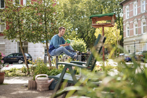 Mature man reading book and sitting on metal structure in garden - KNSF09928