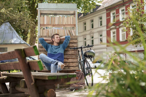 Nachdenklicher Mann lehnt an einem Schrank mit Büchern in der Nähe eines Fahrrads - KNSF09930