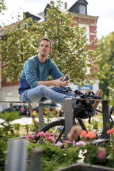 Happy man holding smart phone and sitting on metal structure near bicycle - KNSF09941