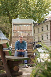 Man using smart phone sitting on bench near cabinet of books - KNSF09945