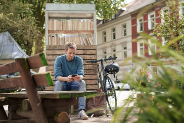 Mature man using smart phone sitting near cabinet of books in suburb - KNSF09946