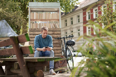 Älterer Mann, der ein Smartphone benutzt, sitzt in der Nähe eines Bücherschranks in einem Vorort - KNSF09946