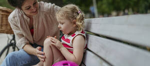 Mother watching little girl blowing on injured knee and putting on band aid - HAPF03426