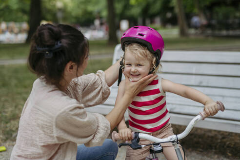 Mother putting safety helmet on little girl's head learning to ride a bike in the city park - HAPF03427
