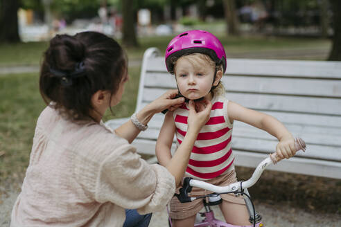 Mother putting safety helmet on little girl's head learning to ride a bike in the city park - HAPF03428