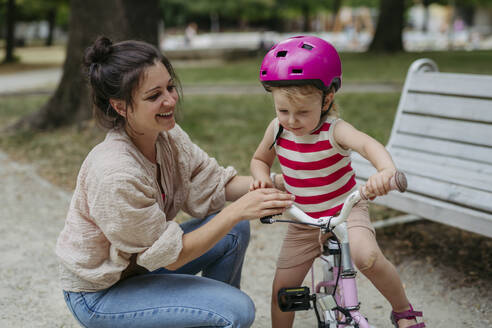 Mother putting safety helmet on little girl's head learning to ride a bike in the city park - HAPF03429