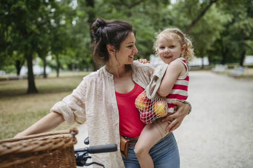 Mother carrying little girl and groceries in gthe city park pushing bicycle - HAPF03432