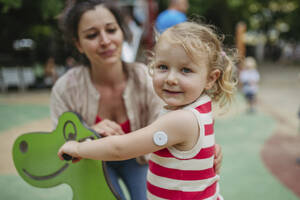 Mother watching little girl with a continuous glucose monitor sitting on dinosaur seesaw on the playground - HAPF03433