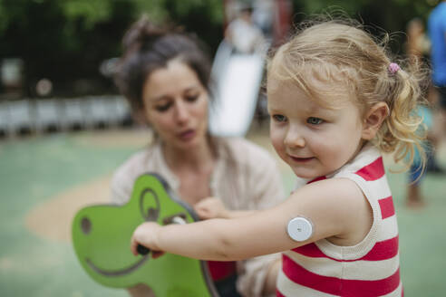 Mother watching little girl with a continuous glucose monitor sitting on dinosaur seesaw on the playground - HAPF03434