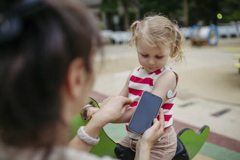 Mother with smartphone checking on little girl with a continuous glucose monitor sensor on her shoulder - HAPF03435