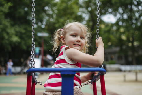 Smiling girl sitting in swing at gthe playground - HAPF03443