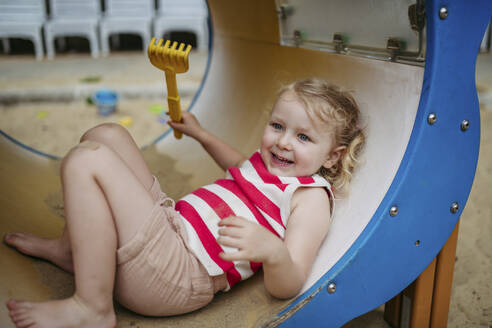 Happy little girl with toy rake lying in climbing frame at the playground - HAPF03448