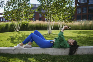 Woman lying on park bench taking selfie listening music after workout - HAPF03479