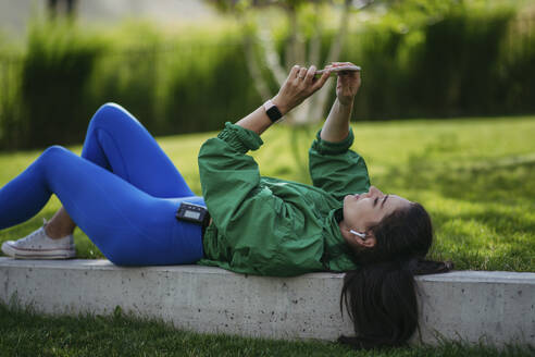 Woman lying on park bench taking selfie listening music after workout - HAPF03480