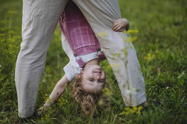 Little girl looking through father's legs holding her upside down - HAPF03493