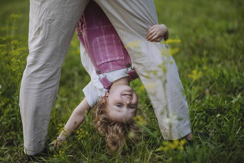 Little girl looking through father's legs holding her upside down - HAPF03493