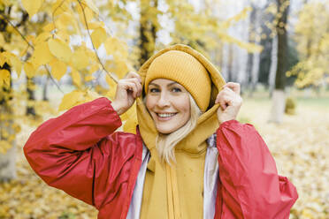 Smiling woman wearing raincoat and knit hat in park - VBUF00487