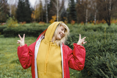 Playful woman wearing raincoat and showing peace sign in park - VBUF00488